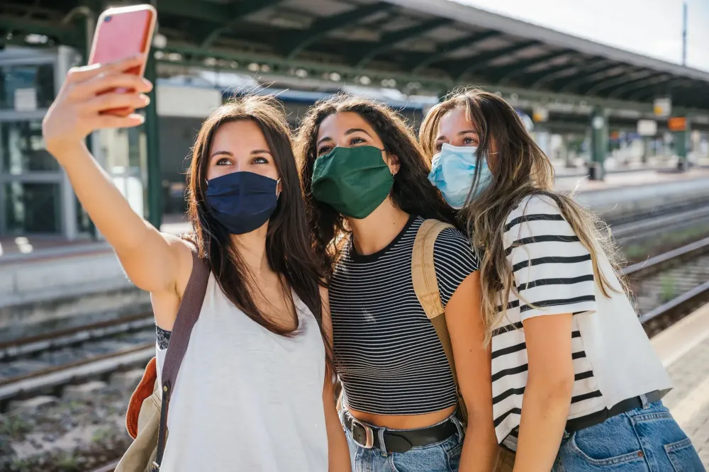 Three young friends taking a selfie in a train station wearing face masks
